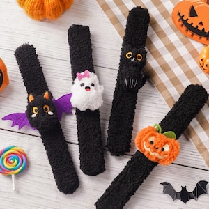 Peut inclure: Quatre bracelets claque noirs et duveteux avec des décorations en peluche sur le thème d'Halloween : une chauve-souris noire aux ailes violettes, un fantôme blanc avec un nœud rose, un chat noir aux yeux jaunes et une citrouille orange.