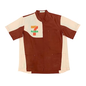 Puede incluir: Camisa de manga corta con diseño de bloques de color en marrón y crema. La camisa tiene cuello mandarín, dos bolsillos delanteros y cierre de cremallera. El logotipo de 7-Eleven está impreso en el pecho.