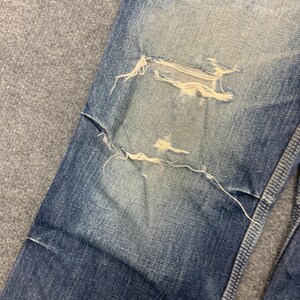 Puede incluir: Primer plano de unos vaqueros de mezclilla azul desgastados. Los vaqueros presentan roturas y desgarros intencionados, con bordes deshilachados visibles. La mezclilla tiene un aspecto descolorido, con parches m&aacute;s claros alrededor de las zonas desgastadas y un tono azul m&aacute;s oscuro en general.