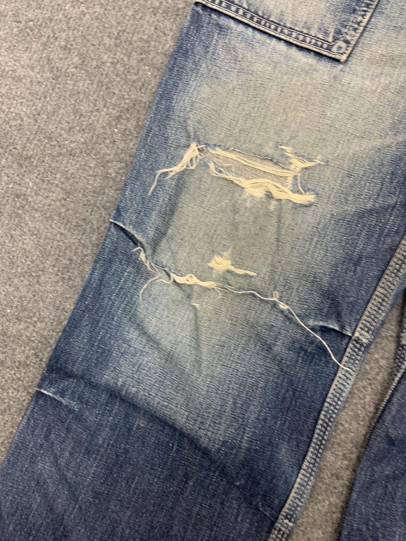 Puede incluir: Primer plano de unos vaqueros de mezclilla azul desgastados. Los vaqueros presentan roturas y desgarros intencionados, con bordes deshilachados visibles. La mezclilla tiene un aspecto descolorido, con parches m&aacute;s claros alrededor de las zonas desgastadas y un tono azul m&aacute;s oscuro en general.