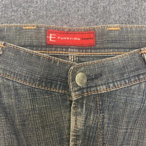 May include: Close-up of denim jeans with a button closure and a red label that reads "E FUNCTION EDWIN". The jeans have a faded blue wash with visible stitching and a button with the letter "E".