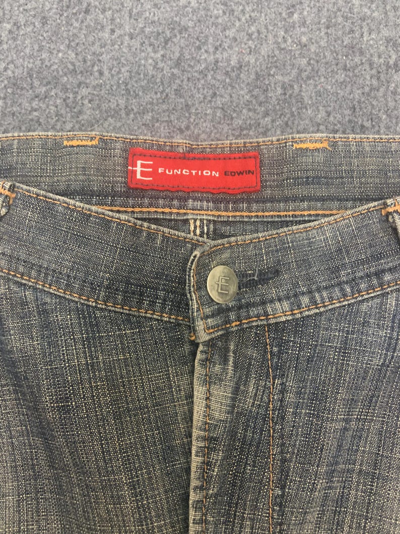 May include: Close-up of denim jeans with a button closure and a red label that reads "E FUNCTION EDWIN". The jeans have a faded blue wash with visible stitching and a button with the letter "E".