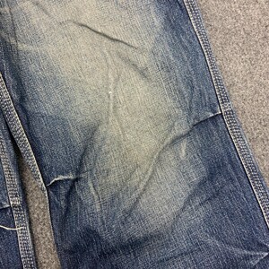 Puede incluir: Primer plano de unos vaqueros azules con desgaste visible. Los vaqueros tienen un aspecto descolorido con parches m&aacute;s claros y costuras blancas contrastantes. La tela muestra signos de uso.