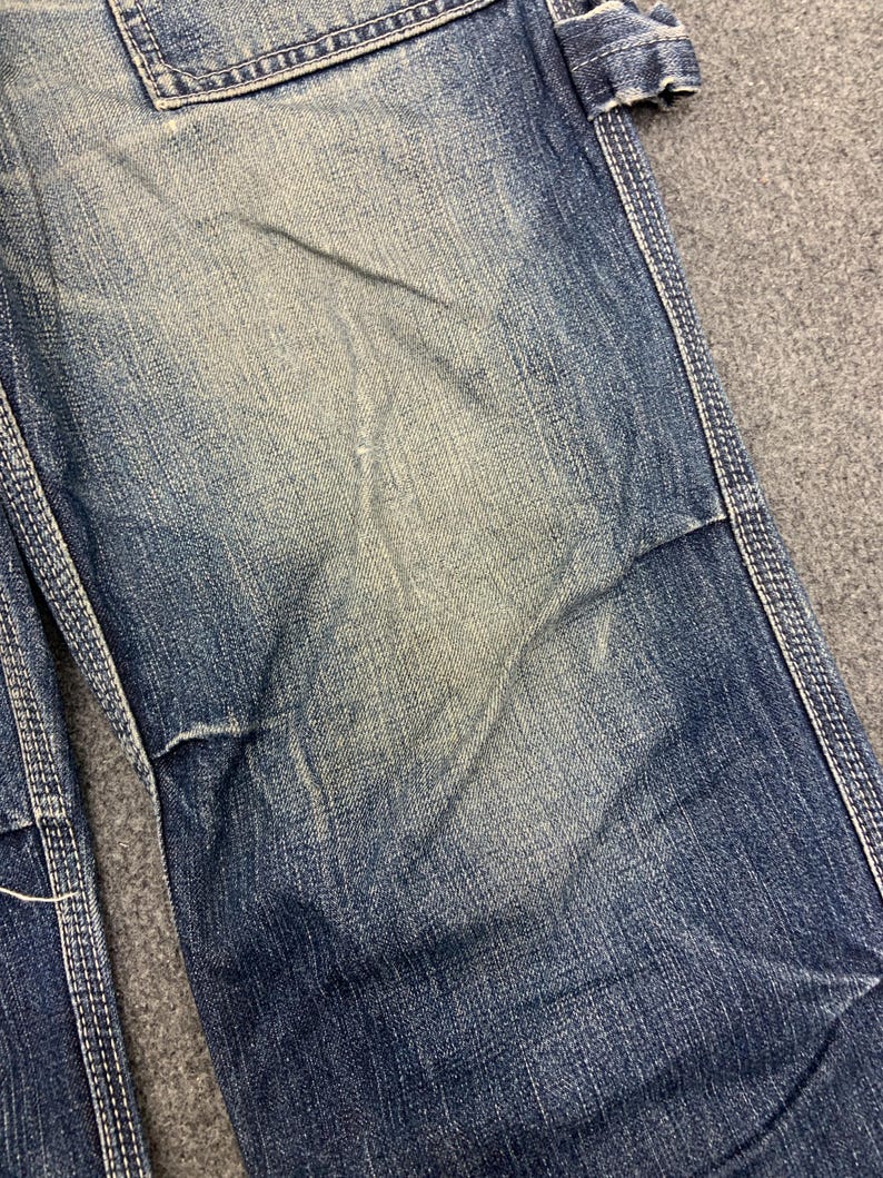 Puede incluir: Primer plano de unos vaqueros azules con desgaste visible. Los vaqueros tienen un aspecto descolorido con parches m&aacute;s claros y costuras blancas contrastantes. La tela muestra signos de uso.