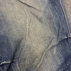 Puede incluir: Primer plano de una secci&oacute;n de vaqueros azules. La tela muestra un efecto de lavado descolorido con &aacute;reas m&aacute;s claras y sombras m&aacute;s oscuras, creando una apariencia texturizada. Costuras visibles a lo largo del borde.