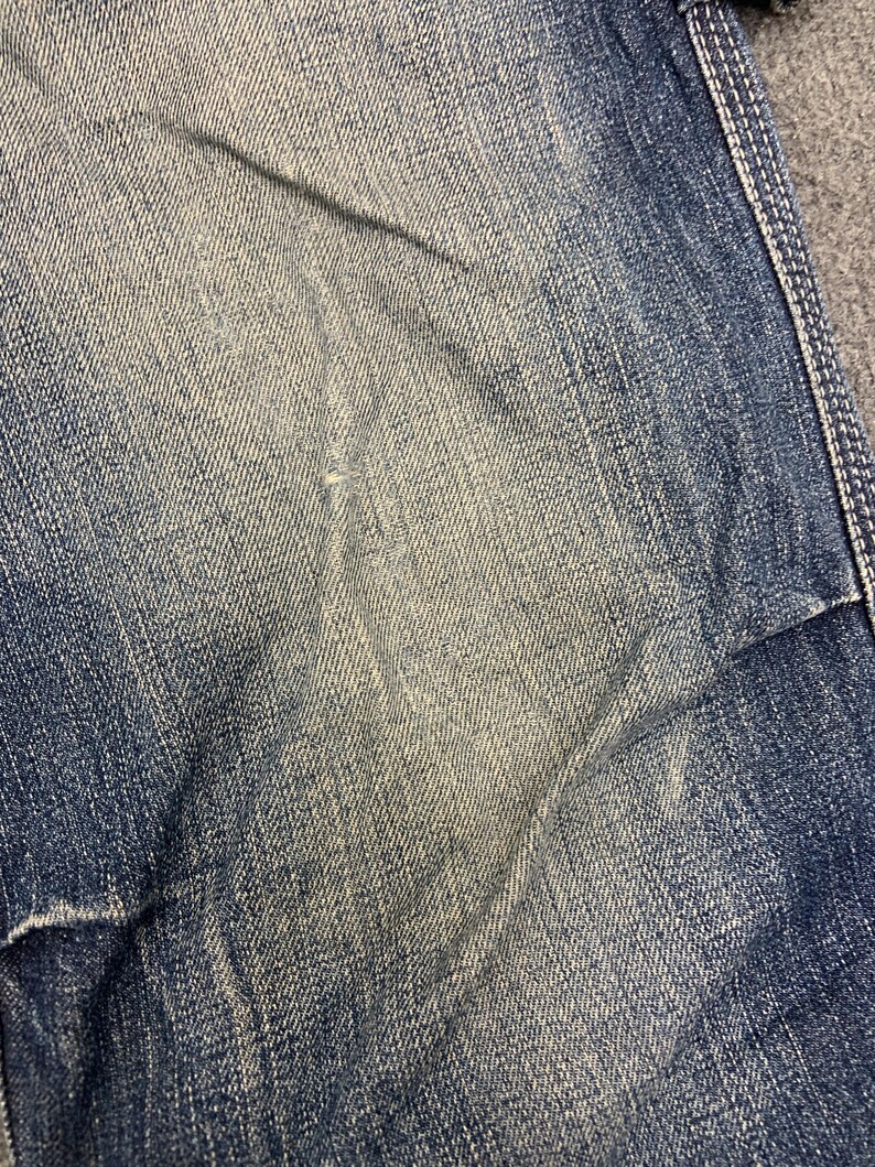 Puede incluir: Primer plano de una secci&oacute;n de vaqueros azules. La tela muestra un efecto de lavado descolorido con &aacute;reas m&aacute;s claras y sombras m&aacute;s oscuras, creando una apariencia texturizada. Costuras visibles a lo largo del borde.