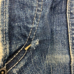 Puede incluir: Primer plano de tejido vaquero azul desgastado. Detalles visibles: costuras blancas, un peque&ntilde;o desgarro y un bot&oacute;n met&aacute;lico. El denim tiene un aspecto descolorido.