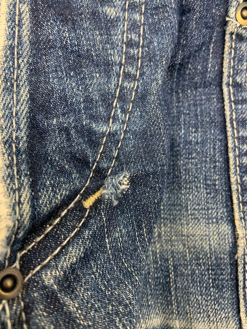 Puede incluir: Primer plano de tejido vaquero azul desgastado. Detalles visibles: costuras blancas, un peque&ntilde;o desgarro y un bot&oacute;n met&aacute;lico. El denim tiene un aspecto descolorido.