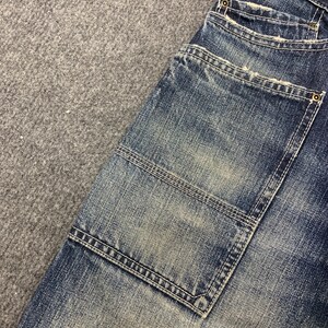 Puede incluir: Primer plano de una secci&oacute;n de vaqueros azules. Los vaqueros tienen un lavado descolorido con desgaste visible. Incluye un bolsillo grande con costuras reforzadas y detalles de botones met&aacute;licos.