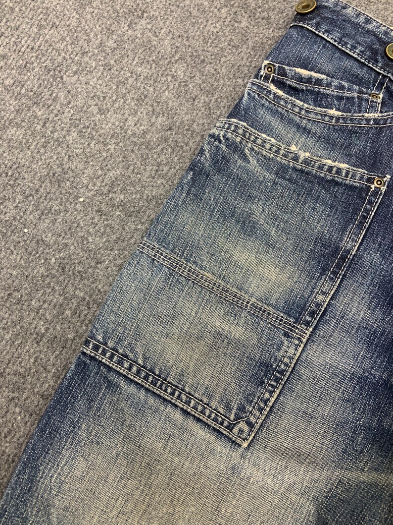 Puede incluir: Primer plano de una secci&oacute;n de vaqueros azules. Los vaqueros tienen un lavado descolorido con desgaste visible. Incluye un bolsillo grande con costuras reforzadas y detalles de botones met&aacute;licos.