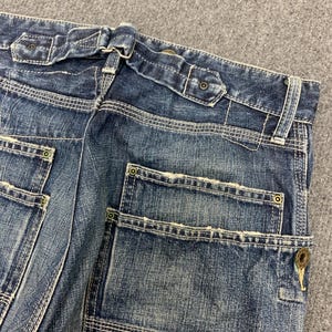 Puede incluir: Primer plano de la parte trasera de unos vaqueros azules. Los vaqueros tienen un dise&ntilde;o &uacute;nico de trabillas y detalles de costura visibles. Los bolsillos cuadrados tienen remaches y cierre de bot&oacute;n. El denim tiene un aspecto descolorido y desgastado.