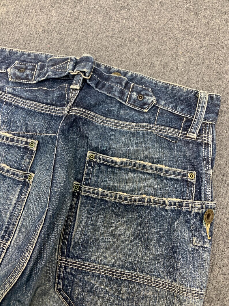 Puede incluir: Primer plano de la parte trasera de unos vaqueros azules. Los vaqueros tienen un dise&ntilde;o &uacute;nico de trabillas y detalles de costura visibles. Los bolsillos cuadrados tienen remaches y cierre de bot&oacute;n. El denim tiene un aspecto descolorido y desgastado.