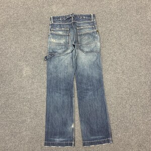 Puede incluir: Vaqueros azules de mezclilla con m&uacute;ltiples bolsillos y un aspecto desgastado. Los vaqueros tienen un corte de pierna recta y cierre de bot&oacute;n y cremallera. Los bolsillos traseros tienen un dise&ntilde;o &uacute;nico. Los vaqueros est&aacute;n planos sobre una superficie gris.