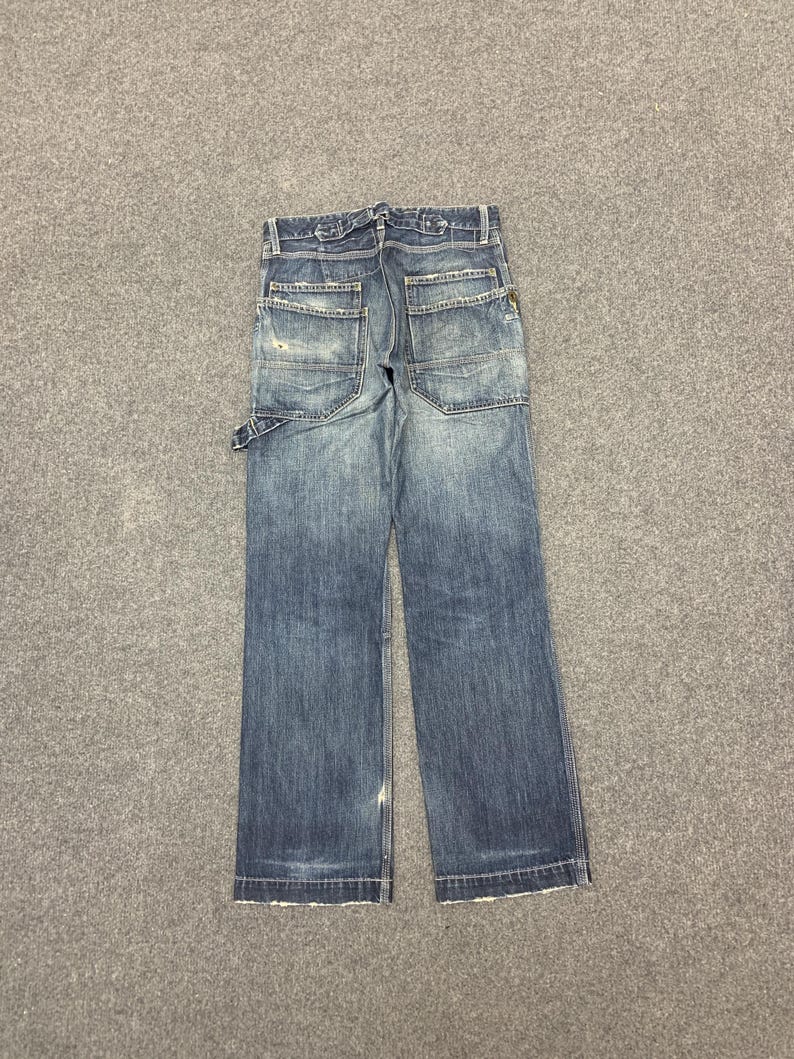 Puede incluir: Vaqueros azules de mezclilla con m&uacute;ltiples bolsillos y un aspecto desgastado. Los vaqueros tienen un corte de pierna recta y cierre de bot&oacute;n y cremallera. Los bolsillos traseros tienen un dise&ntilde;o &uacute;nico. Los vaqueros est&aacute;n planos sobre una superficie gris.