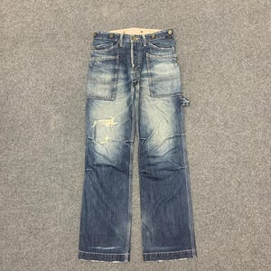 Puede incluir: Vaqueros de mezclilla azul descolorido con m&uacute;ltiples bolsillos y detalles desgastados. Los vaqueros tienen bragueta con botones y un corte relajado. La mezclilla tiene un aspecto desgastado con desgaste visible, d&aacute;ndoles un aspecto vintage.