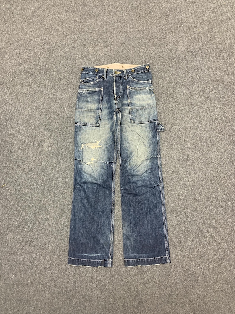 Puede incluir: Vaqueros de mezclilla azul descolorido con m&uacute;ltiples bolsillos y detalles desgastados. Los vaqueros tienen bragueta con botones y un corte relajado. La mezclilla tiene un aspecto desgastado con desgaste visible, d&aacute;ndoles un aspecto vintage.