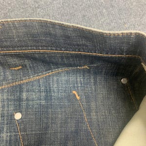 May include: Close-up of a section of blue denim jeans. The jeans have a classic design with visible stitching in a contrasting tan color. Silver metal rivets and a pocket lining are also visible.