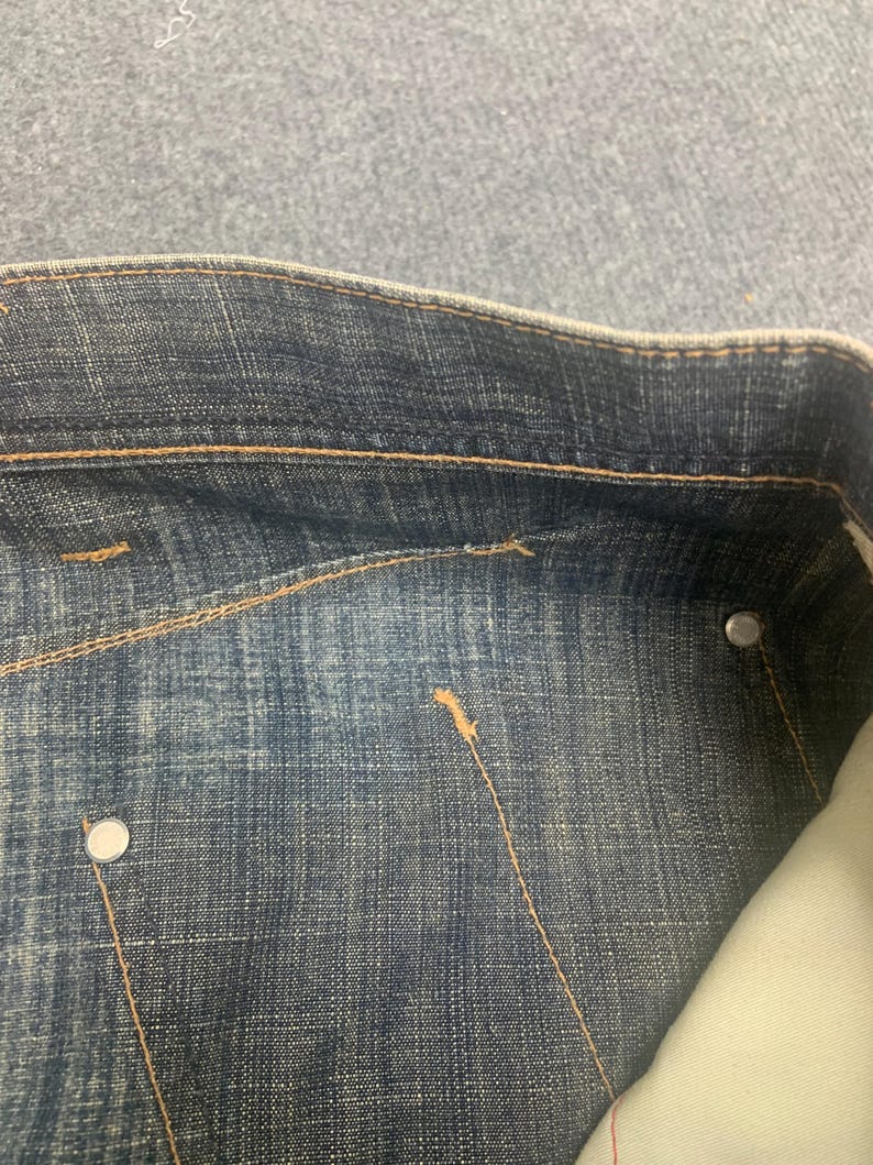 May include: Close-up of a section of blue denim jeans. The jeans have a classic design with visible stitching in a contrasting tan color. Silver metal rivets and a pocket lining are also visible.