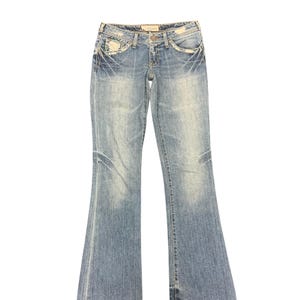 May include: Light blue denim jeans with a faded wash and flared leg. The jeans feature decorative stitching and embellishments near the pockets and along the front. The jeans have a button and zipper closure.