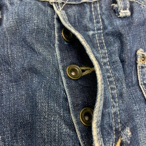 Puede incluir: Primer plano de unos vaqueros de mezclilla azul con costuras visibles y botones de cierre de lat&oacute;n. Los vaqueros muestran signos de desgaste y decoloraci&oacute;n, t&iacute;picos de la mezclilla. La bragueta de botones est&aacute; parcialmente abierta.