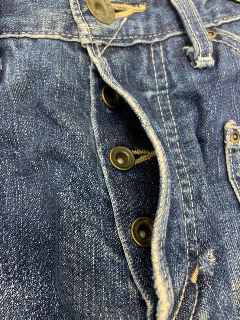 Puede incluir: Primer plano de unos vaqueros de mezclilla azul con costuras visibles y botones de cierre de lat&oacute;n. Los vaqueros muestran signos de desgaste y decoloraci&oacute;n, t&iacute;picos de la mezclilla. La bragueta de botones est&aacute; parcialmente abierta.