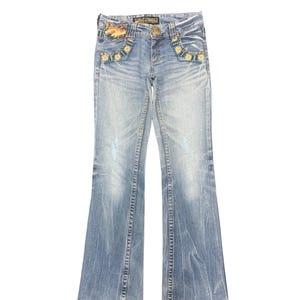 May include: Light blue denim jeans with a flared leg. The jeans feature decorative floral embroidery around the front pockets and a gold-colored button. Faded areas and distressed details add to the vintage look of the jeans.