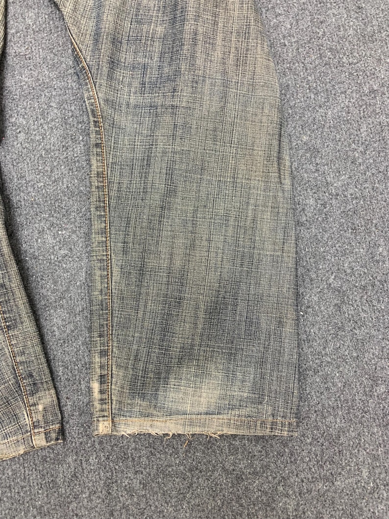 May include: Close-up of faded blue denim jeans. The jeans show wear and tear, with a distressed look. The hem is frayed, and the fabric has a textured appearance. The jeans are likely vintage or have a worn-in style.