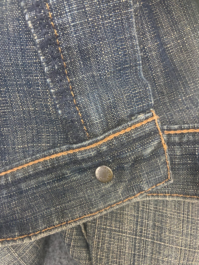 May include: Close-up of a denim jacket. The jacket is a dark blue color with visible stitching in a contrasting tan color. A silver button is visible on the jacket's front. The denim fabric has a textured appearance.