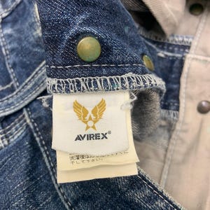 Puede incluir: Primer plano de una chaqueta vaquera con botones color lat&oacute;n. La chaqueta presenta la etiqueta de la marca "AVIREX" con un logotipo de &aacute;guila dorada. La tela es un lavado azul oscuro con costuras visibles y forro beige claro.