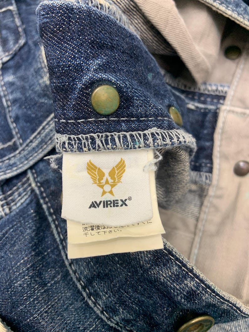 Puede incluir: Primer plano de una chaqueta vaquera con botones color lat&oacute;n. La chaqueta presenta la etiqueta de la marca "AVIREX" con un logotipo de &aacute;guila dorada. La tela es un lavado azul oscuro con costuras visibles y forro beige claro.
