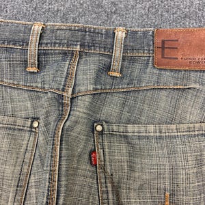 May include: Close-up of the back of blue denim jeans. The jeans have a brown leather patch with the letter "E" and the brand name "FUNCTION EDWIN". The back pockets have rivets and the Edwin red tag. The stitching is in a contrasting orange color.