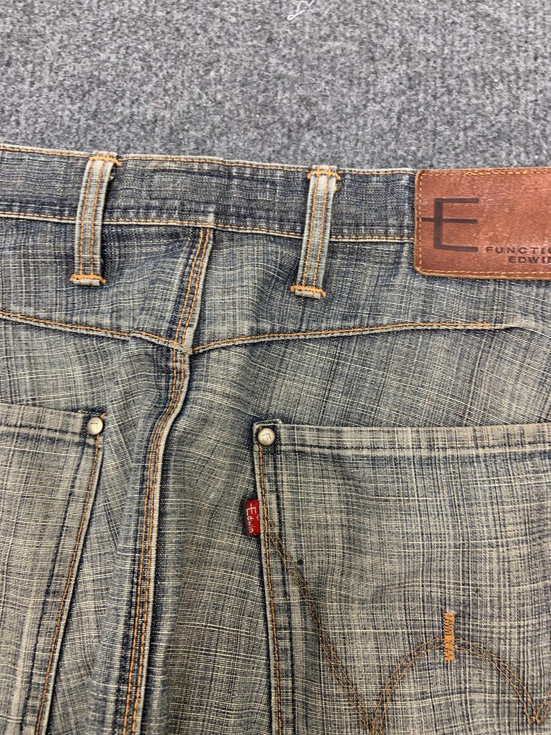 May include: Close-up of the back of blue denim jeans. The jeans have a brown leather patch with the letter "E" and the brand name "FUNCTION EDWIN". The back pockets have rivets and the Edwin red tag. The stitching is in a contrasting orange color.