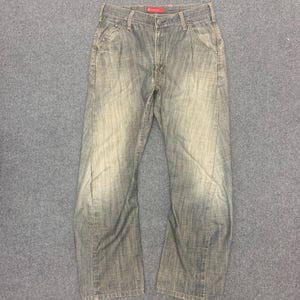 May include: Faded blue denim jeans with a classic five-pocket design. The jeans have a straight leg cut and a worn appearance, with subtle fading and distressing. The waistband features a red tag. The jeans are laid flat on a gray surface.