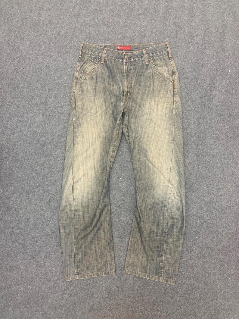 May include: Faded blue denim jeans with a classic five-pocket design. The jeans have a straight leg cut and a worn appearance, with subtle fading and distressing. The waistband features a red tag. The jeans are laid flat on a gray surface.
