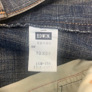 May include: Close-up of a pair of blue denim jeans. The tag reads "EDWIN" and "E2000". The size is 33x33 inches. The jeans are made of a durable denim fabric.