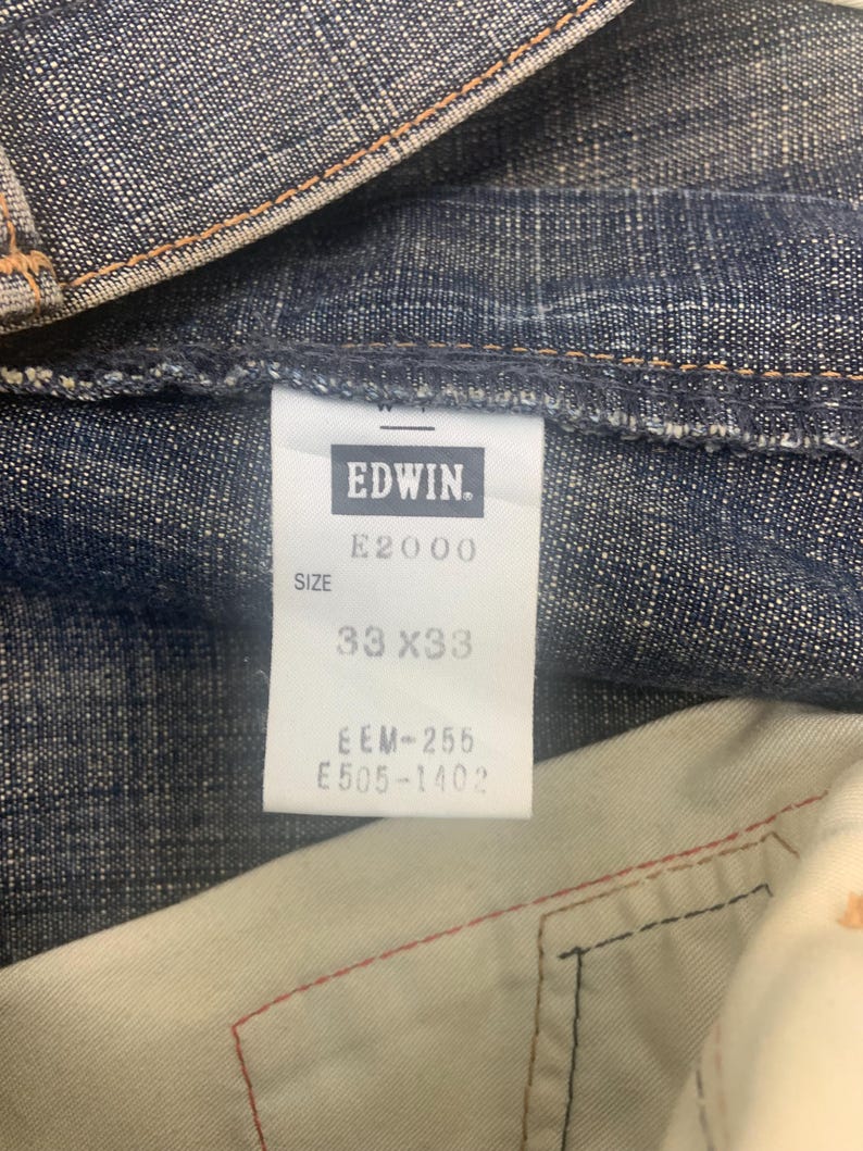 May include: Close-up of a pair of blue denim jeans. The tag reads "EDWIN" and "E2000". The size is 33x33 inches. The jeans are made of a durable denim fabric.