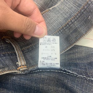 May include: Close-up of denim fabric with a tag. The tag has Japanese text and washing instructions. The fabric is a medium blue color with visible stitching and a textured appearance. The tag indicates the fabric is 70% cotton and 30% polyester.