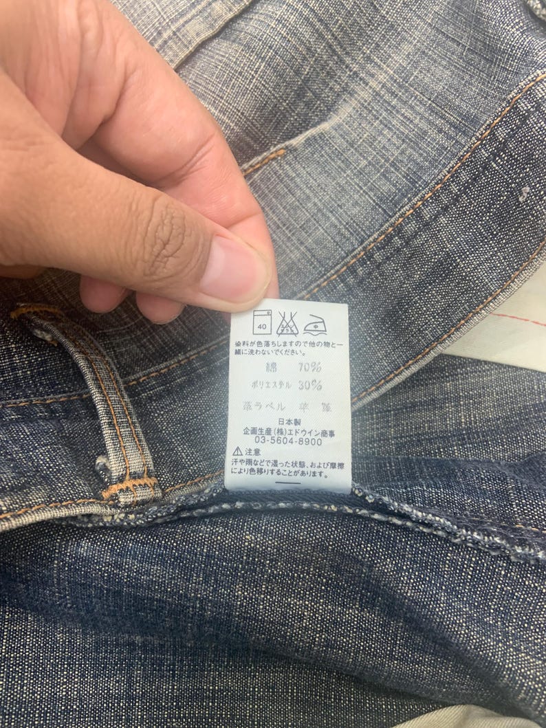 May include: Close-up of denim fabric with a tag. The tag has Japanese text and washing instructions. The fabric is a medium blue color with visible stitching and a textured appearance. The tag indicates the fabric is 70% cotton and 30% polyester.