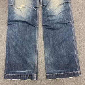 Puede incluir: Primer plano de unos vaqueros de mezclilla azules con detalles desgastados. Los vaqueros tienen un lavado descolorido, una rodilla rasgada y un corte recto. Las costuras son visibles a lo largo de las costuras y el dobladillo est&aacute; deshilachado.