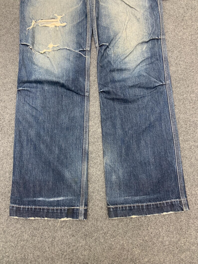 Puede incluir: Primer plano de unos vaqueros de mezclilla azules con detalles desgastados. Los vaqueros tienen un lavado descolorido, una rodilla rasgada y un corte recto. Las costuras son visibles a lo largo de las costuras y el dobladillo est&aacute; deshilachado.