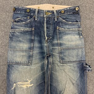 Puede incluir: Pantalones de trabajo vaqueros azules descoloridos con m&uacute;ltiples bolsillos y detalles desgastados. Los pantalones tienen bragueta con botones, costuras reforzadas y cinturilla de color claro. El vaquero tiene un aspecto vintage desgastado.