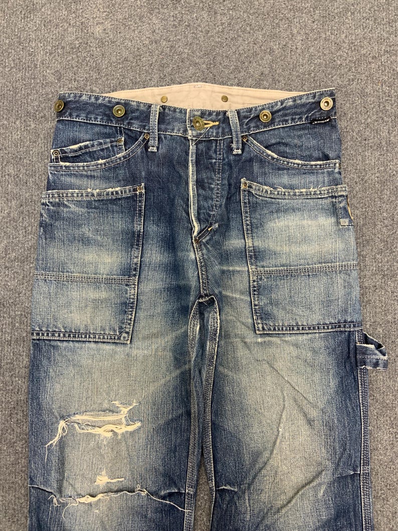Puede incluir: Pantalones de trabajo vaqueros azules descoloridos con m&uacute;ltiples bolsillos y detalles desgastados. Los pantalones tienen bragueta con botones, costuras reforzadas y cinturilla de color claro. El vaquero tiene un aspecto vintage desgastado.