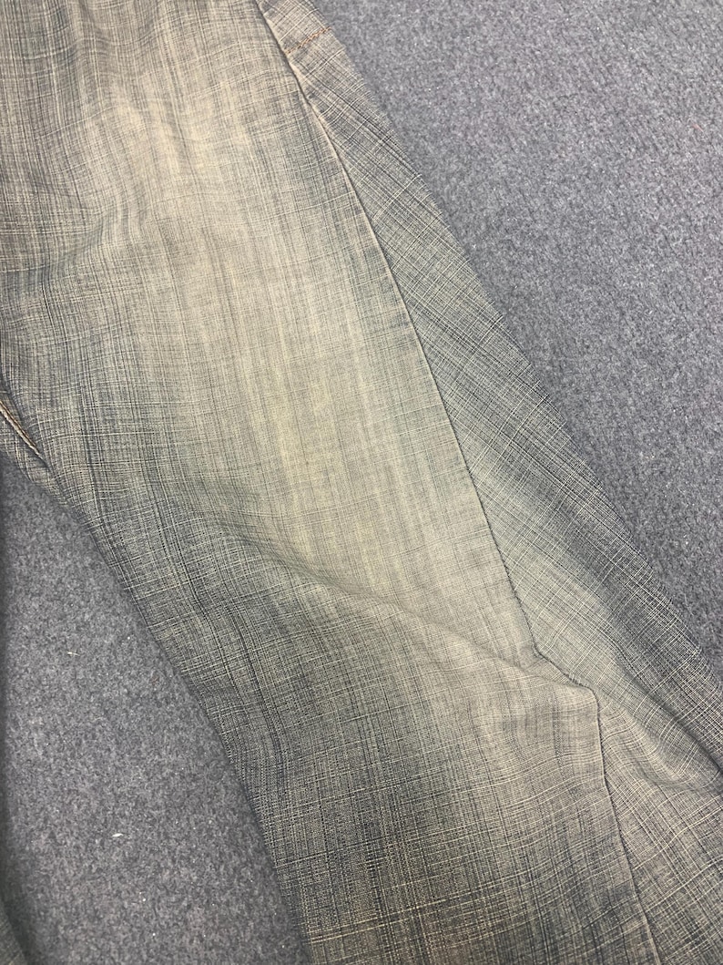 May include: Close-up of denim pants in shades of gray and beige. The fabric has a textured appearance with visible vertical and horizontal threads. The pants are folded, showing a seam and a portion of the leg.