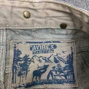 Puede incluir: Primer plano del cuello de una chaqueta vaquera con una etiqueta. La etiqueta presenta el texto "AVIREX CAMPING" sobre un gr&aacute;fico azul de un ciervo, &aacute;rboles y monta&ntilde;as. Se ven botones de color lat&oacute;n.