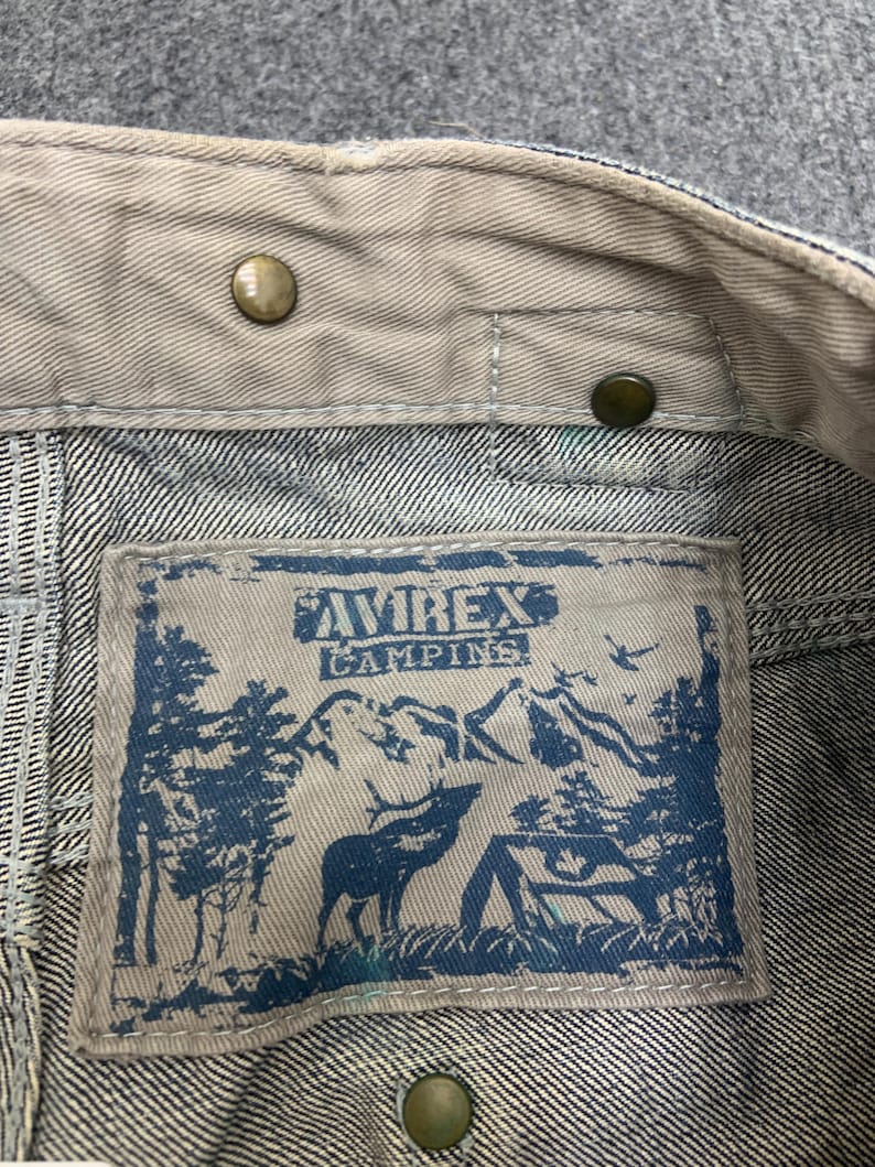 Puede incluir: Primer plano del cuello de una chaqueta vaquera con una etiqueta. La etiqueta presenta el texto "AVIREX CAMPING" sobre un gr&aacute;fico azul de un ciervo, &aacute;rboles y monta&ntilde;as. Se ven botones de color lat&oacute;n.