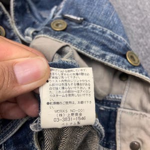 Puede incluir: Primer plano de una chaqueta vaquera azul con botones color lat&oacute;n. El forro interior de la chaqueta es visible, junto con una etiqueta con texto japon&eacute;s y la marca "WORKS NO-001".