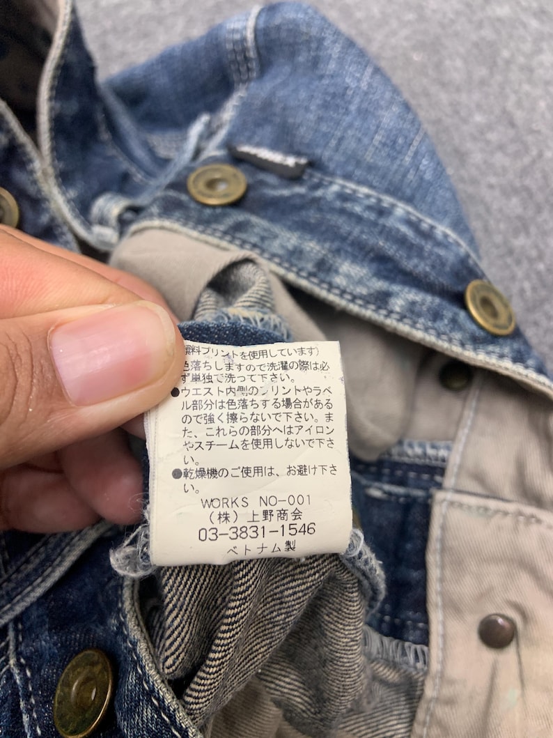 Puede incluir: Primer plano de una chaqueta vaquera azul con botones color lat&oacute;n. El forro interior de la chaqueta es visible, junto con una etiqueta con texto japon&eacute;s y la marca "WORKS NO-001".