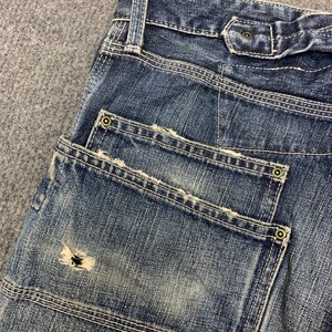 Puede incluir: Primer plano de unos vaqueros azules con costuras visibles y detalles desgastados. Los vaqueros tienen dos bolsillos traseros con remaches decorativos y una zona desgastada en un bolsillo. El tejido vaquero tiene un aspecto descolorido.