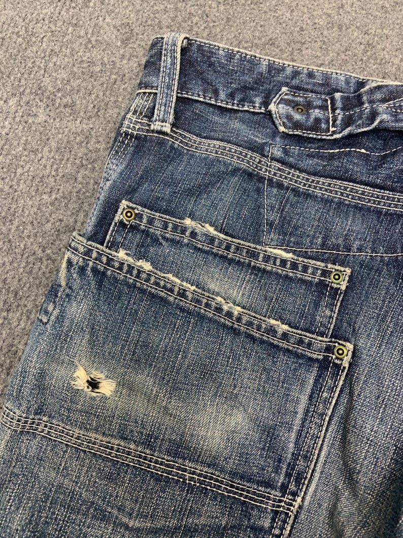 Puede incluir: Primer plano de unos vaqueros azules con costuras visibles y detalles desgastados. Los vaqueros tienen dos bolsillos traseros con remaches decorativos y una zona desgastada en un bolsillo. El tejido vaquero tiene un aspecto descolorido.