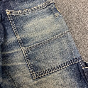 Puede incluir: Primer plano de una secci&oacute;n de vaqueros azules. Los vaqueros tienen un bolsillo grande y cuadrado con costuras reforzadas. El tejido vaquero muestra signos de desgaste y decoloraci&oacute;n, con zonas m&aacute;s claras que contrastan con el tejido azul m&aacute;s oscuro. Se ve un peque&ntilde;o remache met&aacute;lico.